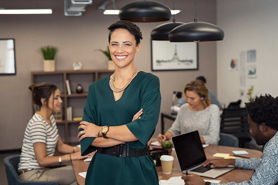 Business Insurance - Portrait of a Smiling Successful Young Business Woman Standing in a Modern Office with Coworkers Blurred in the Background