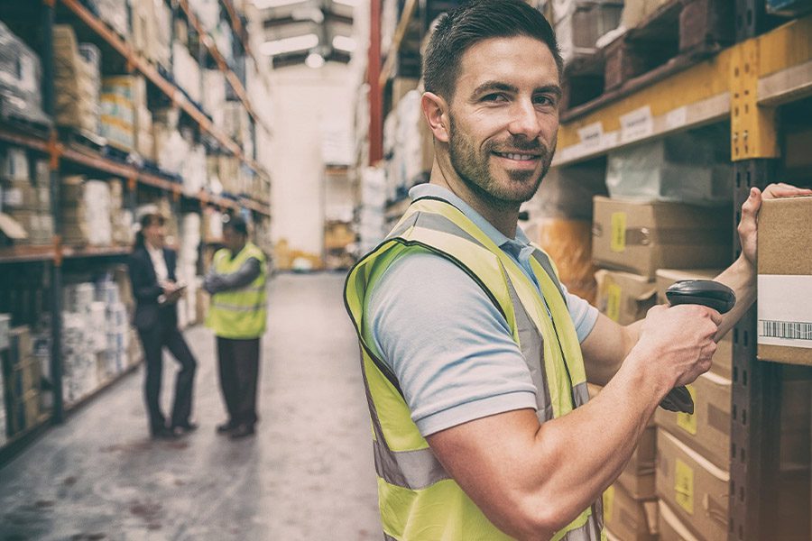 Warehousing-and-Logistics-Insurance-Portrait-of-a-Warehouse-Worker-Scanning-a-Box-While-Smiling-With-Fellow-Colleagues-Blurred-in-the-Distance